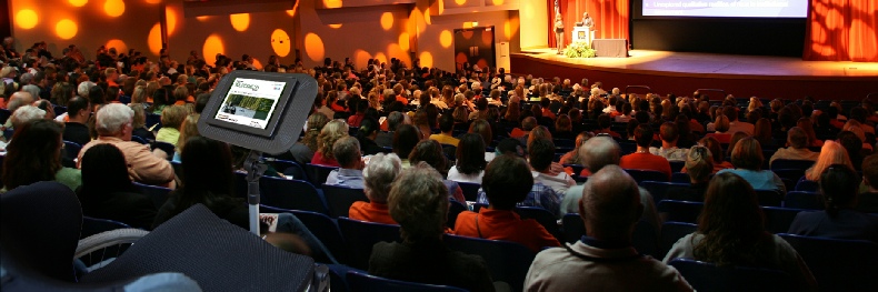 Photo of wheelchair with multi-table attachment with a tablet.  Background image shows a conference